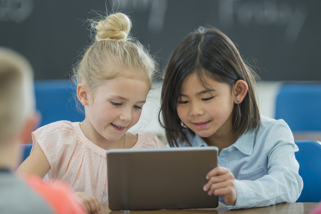 A multi-ethnic group of elementary age school children look at a digital tablet together. Young blonde Caucasian girl is wearing a pink top and young Asian brunette girl is wearing a blue button-up blouse.