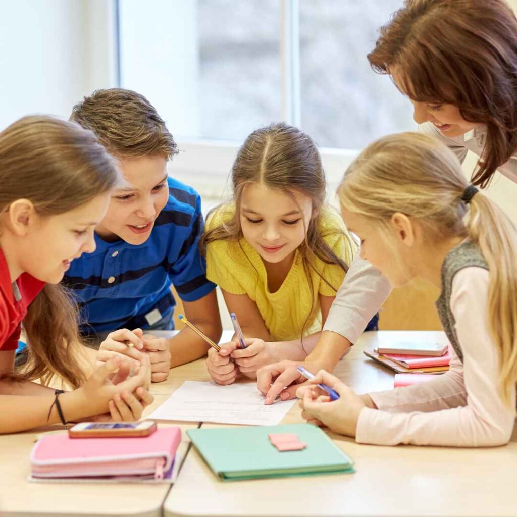 Four children gathered around a desk with a teacher at school.