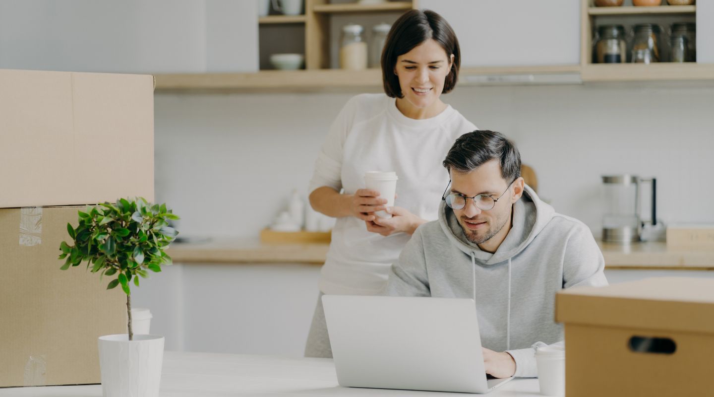 couple sat at a counter top, with moving boxes, looking at their laptop