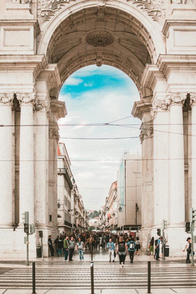 A large stone archway in Portugal leading to a bustling pedestrian street filled with people. The sky is partly cloudy.