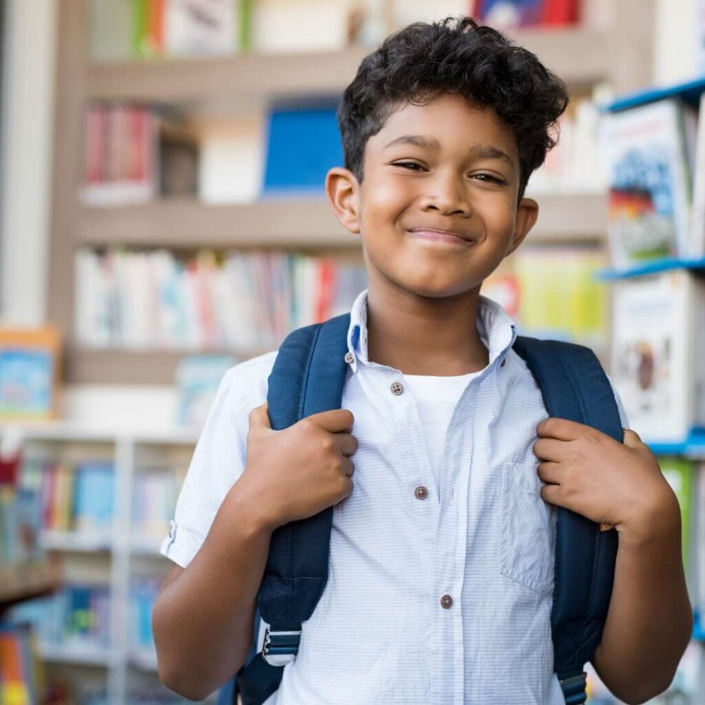 A young boy with a backpack smiles confidently in a school library, ready for a new learning adventure. Santa Fe Relocation supports families in finding the right schools and settling into their new home abroad.