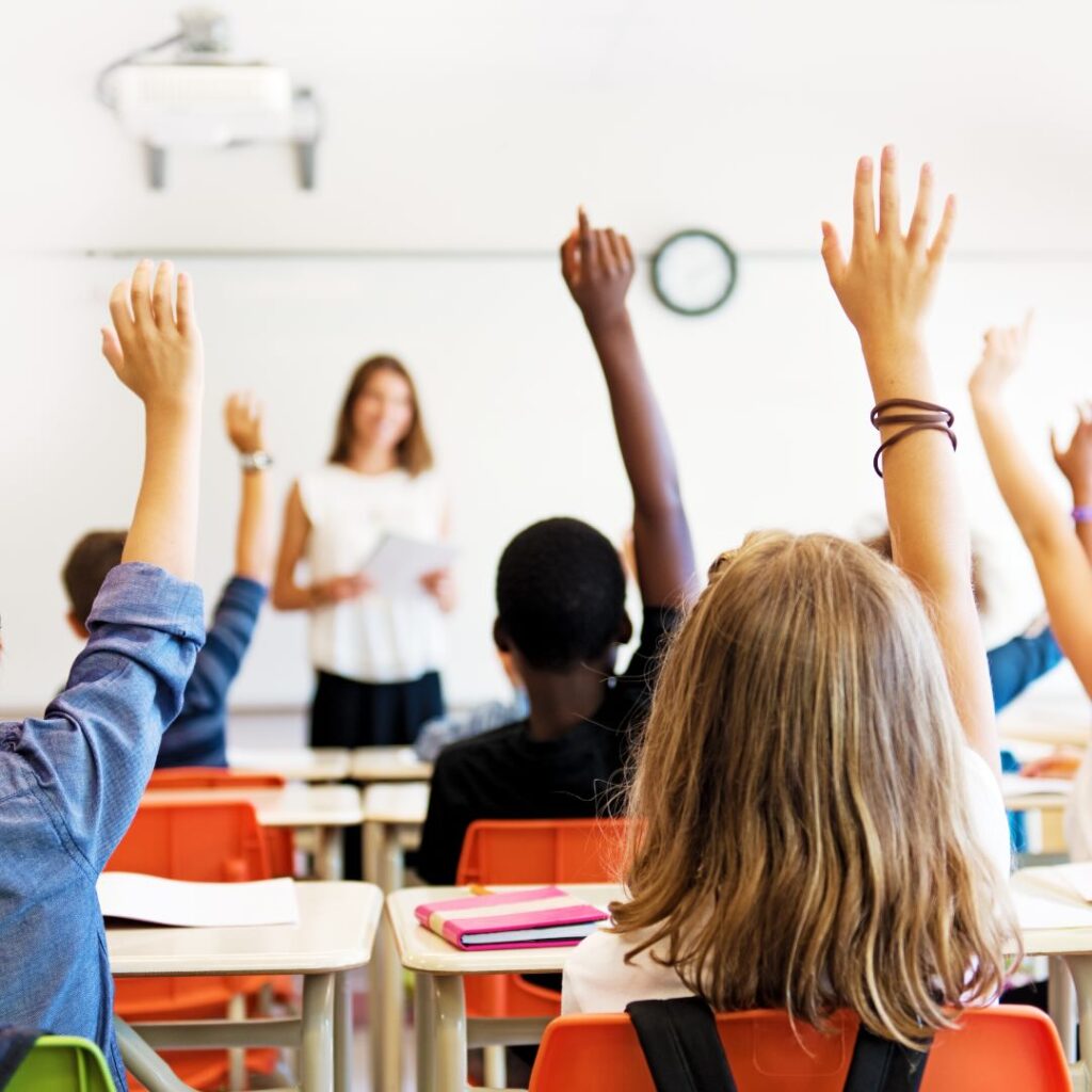 A diverse group of young students eagerly raise their hands in a bright and engaging classroom while a teacher stands at the front. Moving internationally with Santa Fe Relocation ensures your children transition smoothly into new schools worldwide.